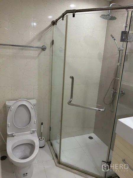 Condominium for rent in Watthana, Bangkok - Bathroom with glass-enclosed rain shower and toilet on light tile walls.