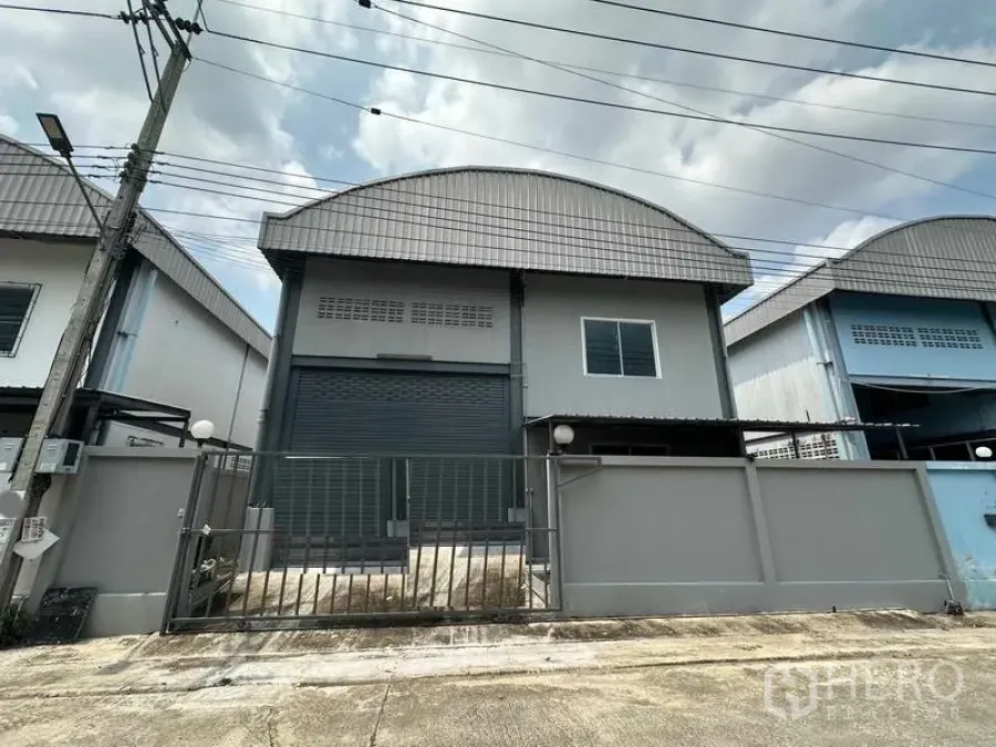 คลังสินค้า ให้เช่าในPak Kret, Nonthaburi - ด้านหน้าโกดังพร้อมรั้วและประตูม้วนขนาดใหญ่ หลังคาโค้งเมทัลชีท