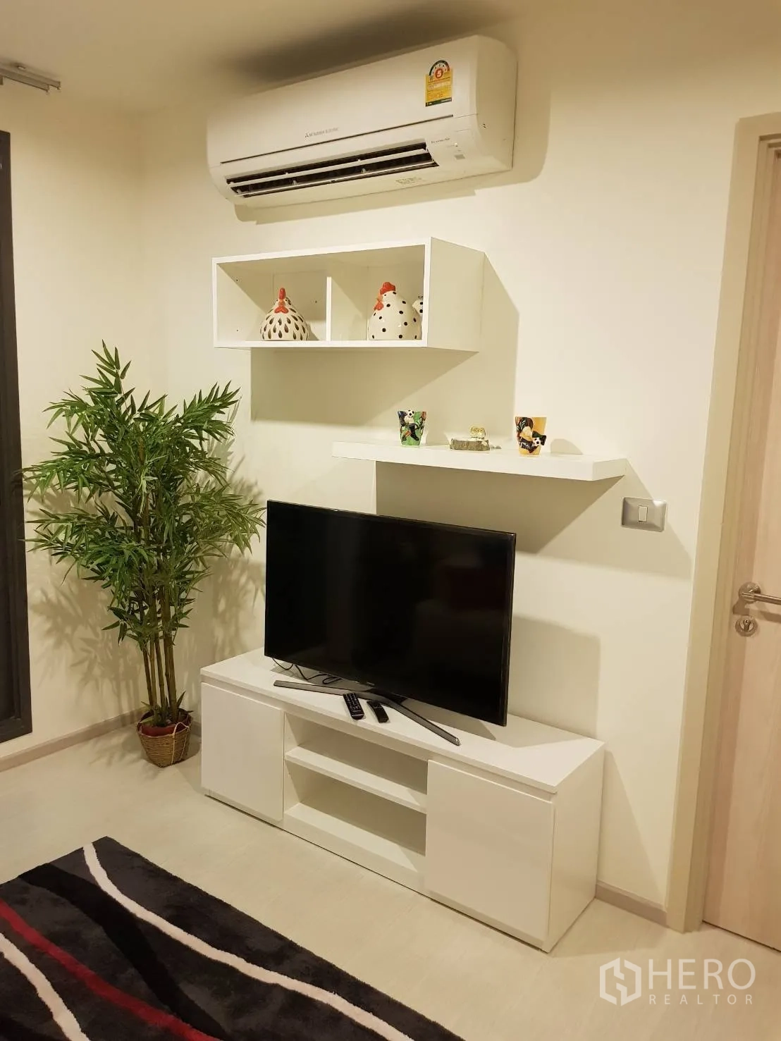 Condominium for rent in Khlong Toei, Bangkok - Media wall with TV, floating shelves, air conditioner, potted plant, and modern rug.