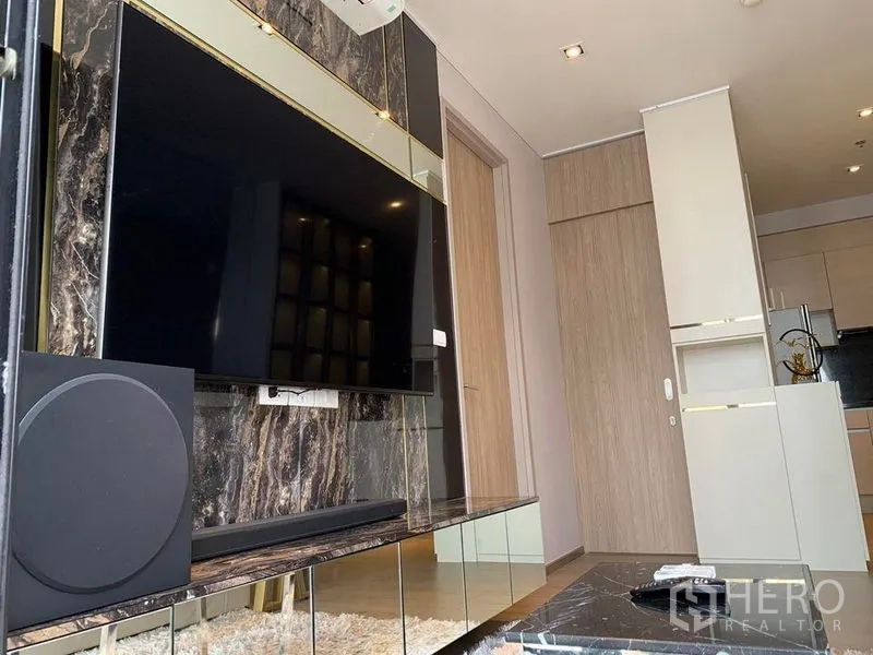 คอนโด ขาย - ผนังทีวีลายหินอ่อนในห้องนั่งเล่น หันหน้าเข้าครัวขนาดกะทัดรัด ในคอนโดกรุงเทพฯ