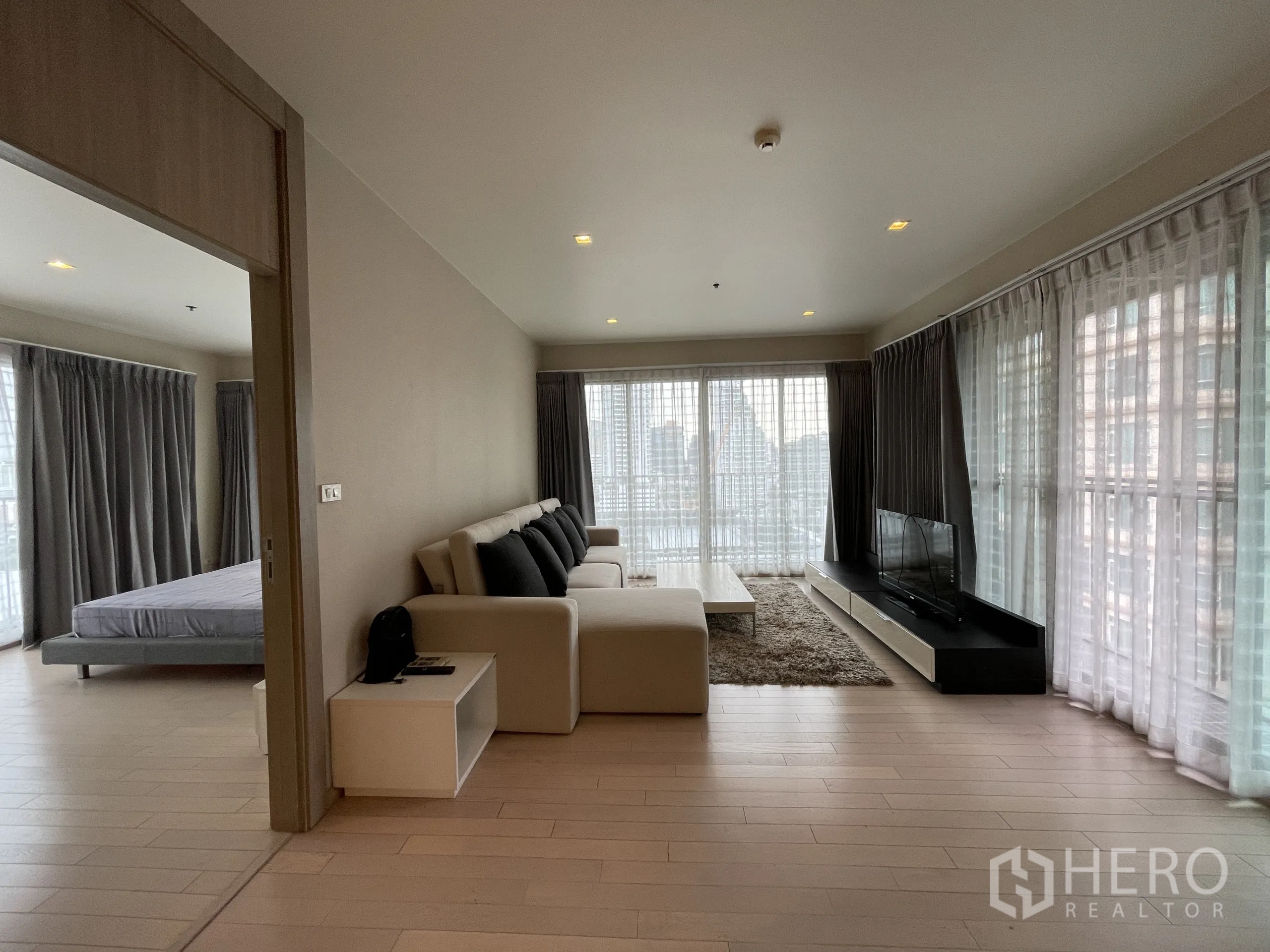 公寓 出租在Watthana, Bangkok - 宽阔起居区，转角沙发与电视，门内可见卧室，周围高窗环绕。