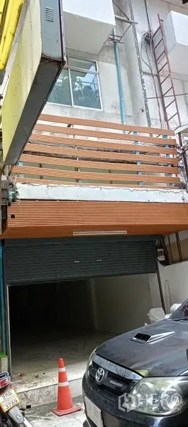 อาคารพาณิชย์ ให้เช่าในWatthana, Bangkok - ด้านหน้าตึกแถวพร้อมระเบียงไม้บังตาและประตูม้วนโลหะเปิดครึ่งหนึ่ง มีนั่งร้านและรถจอดด้านหน้า