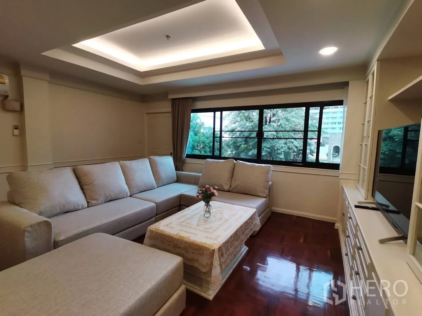 Condominium for rent in Watthana, Bangkok - Living room with large windows, L-shaped sofa, coffee table, TV unit, and a leafy outlook.