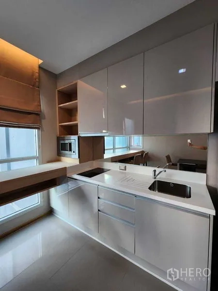 Condominium for sale in Ratchathewi, Bangkok - Ratchathewi condo kitchen with glossy cabinets, cooktop, sink and a breakfast bar beside large windows.