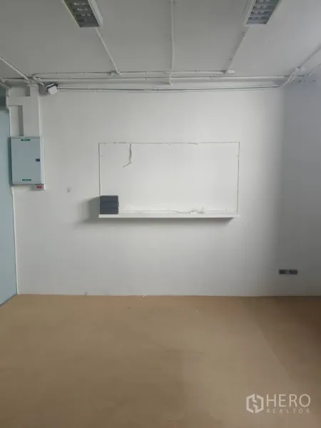 Office Space for rent in Khlong Toei, Bangkok - Minimal white office room with tan flooring, a wall shelf, exposed ceiling conduits and fluorescent lights.