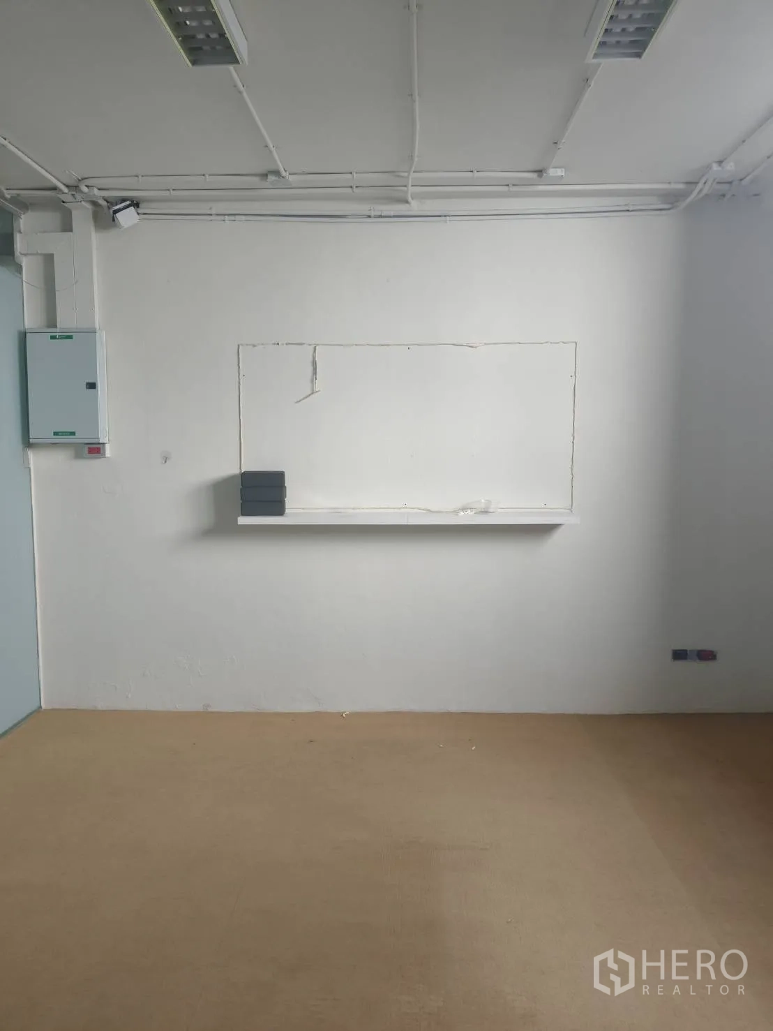 Office Space for rent in Khlong Toei, Bangkok - Minimal white office room with tan flooring, a wall shelf, exposed ceiling conduits and fluorescent lights.