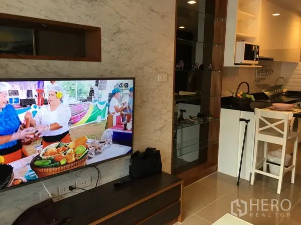 Condominium for rent in Watthana, Bangkok - Open-plan living area with wall-mounted TV facing a compact kitchen with breakfast bar and glass shelving.