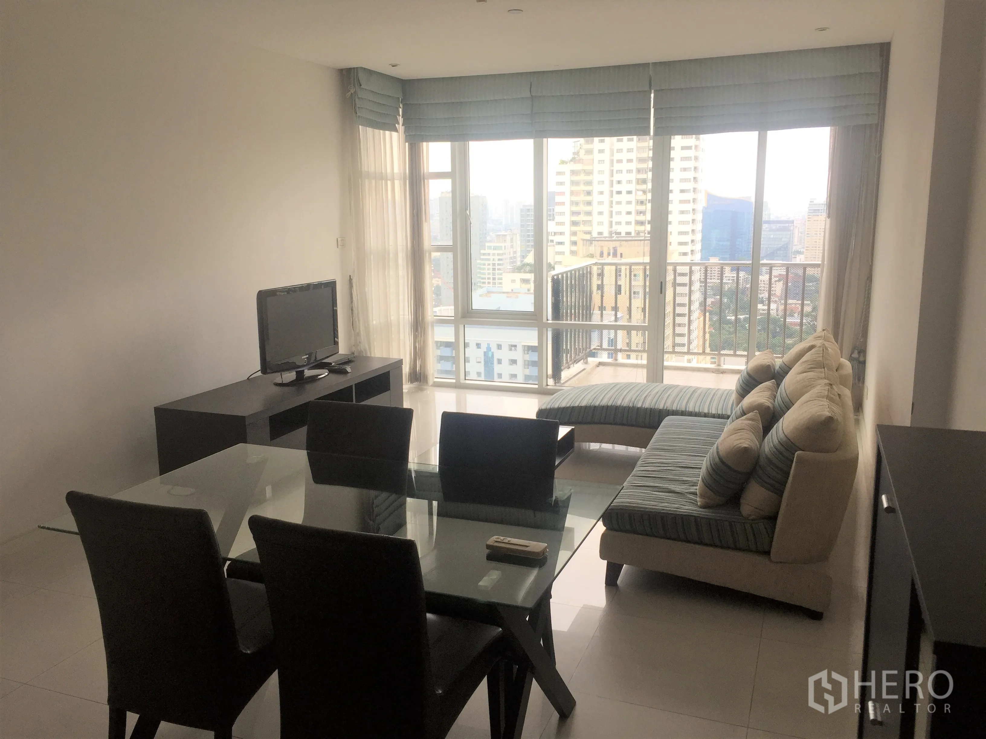 Condominium for rent or sale in Watthana, Bangkok - Bright living and dining area with glass table, sofa, TV console and floor-to-ceiling windows opening to a city-view balcony.