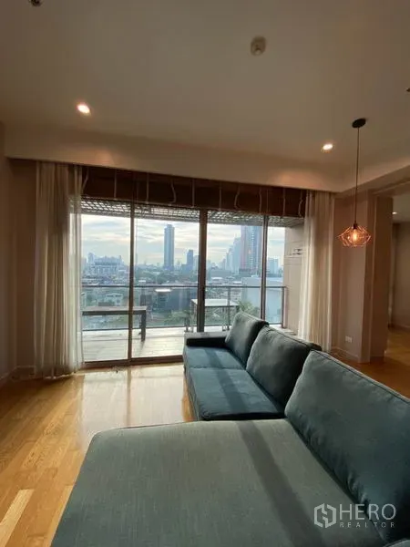Condominium for rent or sale in Yan Nawa, Bangkok - Floor-to-ceiling sliding doors open to a balcony with city skyline views behind a long blue sofa.