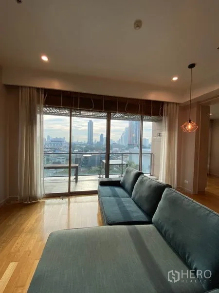 Condominium for rent or sale in Yan Nawa, Bangkok - Floor-to-ceiling sliding doors open to a balcony with city skyline views behind a long blue sofa.