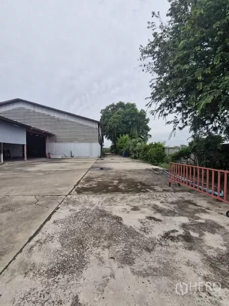 货栈 出租在Lam Luk Ka, Pathum Thani - 仓库侧面混凝土院坝，旁有金属板墙与绿化树木