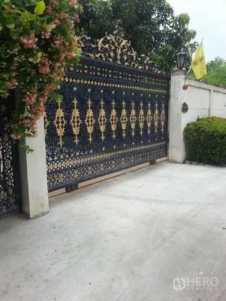 独立式住宅 出租在Bang Na, Bangkok - 带金色装饰的锻铁滑动大门与入口，一侧为修剪的灌木。