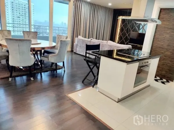 Condominium for rent in Khlong San, Bangkok - Open‑plan living and dining area with a kitchen island, round table, and floor‑to‑ceiling windows leading to a balcony with skyline views.