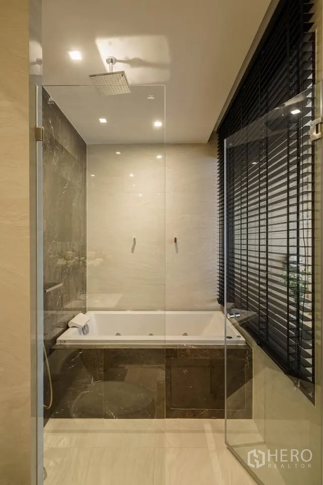 Condominium for rent in Bang Rak, Bangkok - Bathroom with glass-enclosed rain shower and marble-trimmed bathtub beside window blinds.
