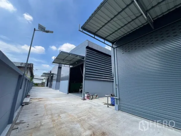 货栈 出租在Prawet, Bangkok - 仓库园区宽阔混凝土车道，多个卷闸门单元与路灯。