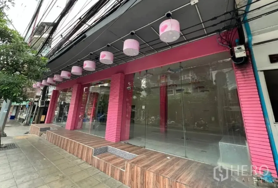 店屋 出租在Watthana, Bangkok - 宽阔玻璃门面，粉色立柱及沿人行道的抬高木质平台。