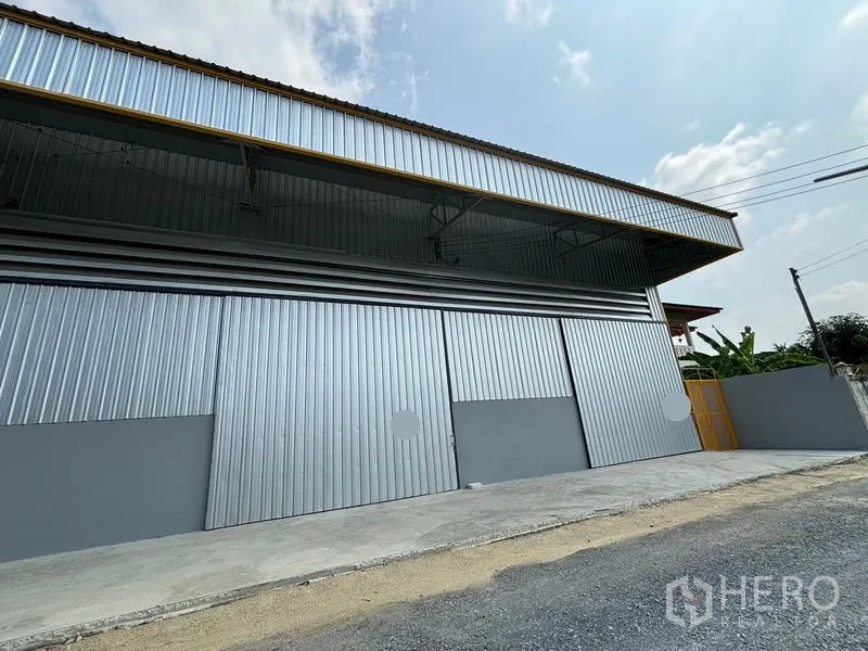 货栈 出租在Mueang Pathum Thani, Pathum Thani - 仓库外立面斜角视图，多扇滑动门与侧门。