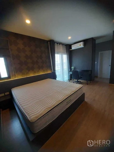 Detached House for rent in Bang Bo, Samut Prakan - Primary bedroom with large bed frame, wood floor, desk and window with sheer curtains.