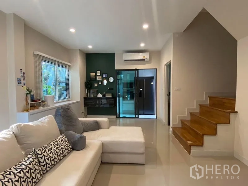 Townhouse for rent in Saphan Sung, Bangkok - Bright open-plan living room with white sectional sofa, wood staircase, recessed lighting and a glass sliding partition with air conditioning.