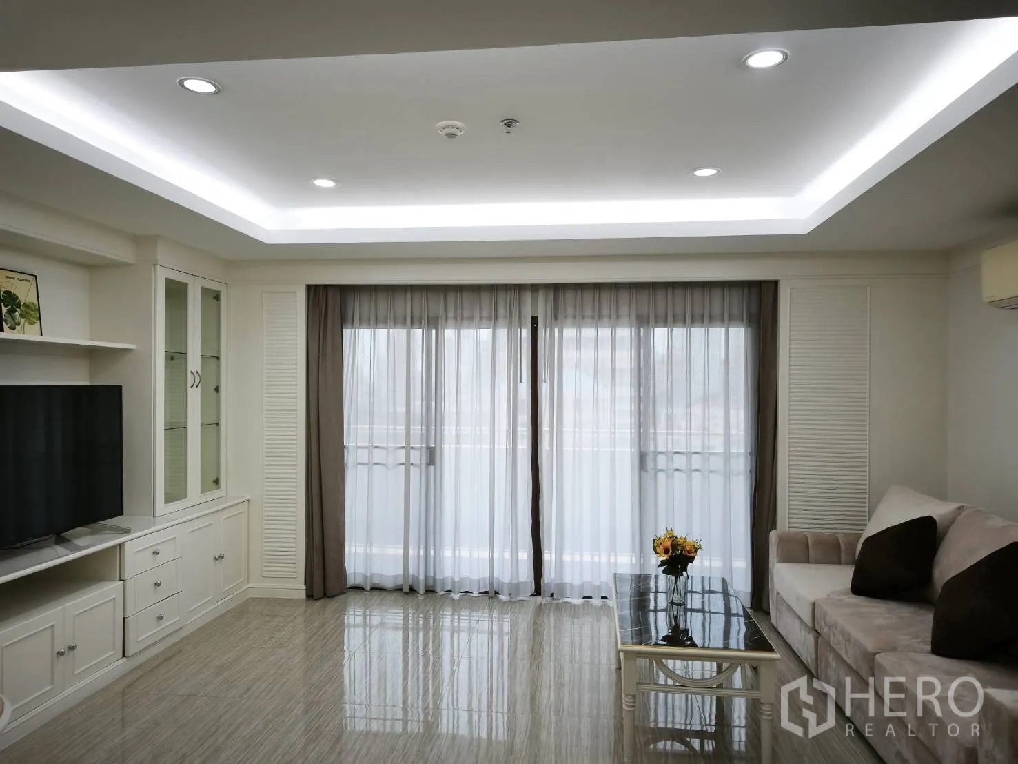 Condominium for rent in Watthana, Bangkok - Living room with built-in TV cabinet, sofa, recessed lighting, and sheer curtains over balcony doors.