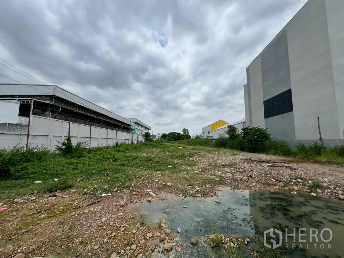 Land for sale - Vacant industrial land with gravel and puddles between large warehouse buildings under a cloudy sky.