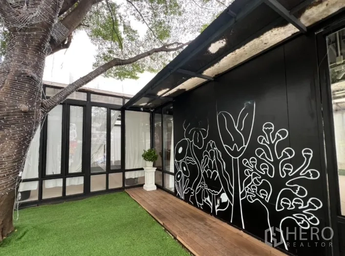 零售空间 出租在Watthana, Bangkok - 黑色立面与花卉壁画的现代商铺，木平台与人造草坪，枝繁叶茂的大树遮荫。