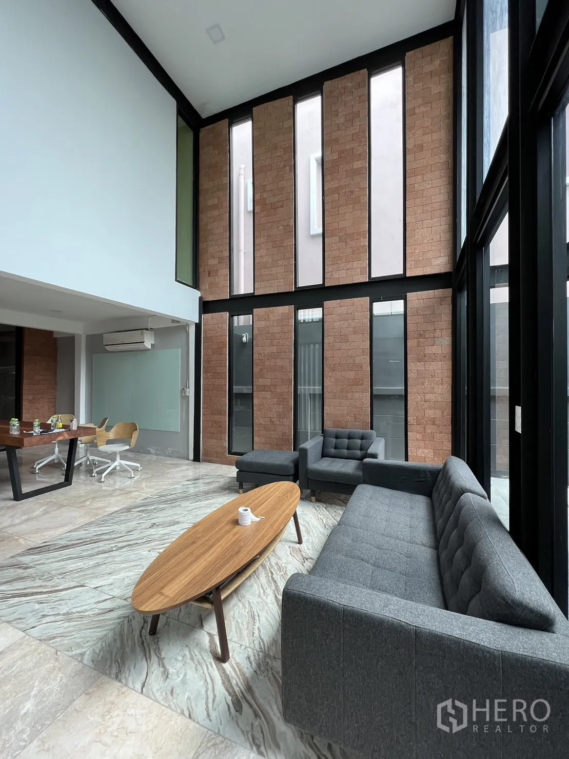 บ้าน ให้เช่าหรือขายในPhra Khanong, Bangkok - ห้องนั่งเล่นเพดานสูงสองชั้น ผนังอิฐโชว์ กระจกเต็มผนัง โซฟาสีเทา และพื้นลายหินอ่อน
