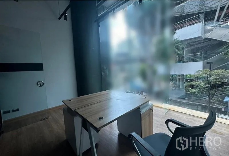 Office Space for rent - Compact office with wooden desk, ergonomic chair and floor-to-ceiling glass overlooking a modern atrium in Watthana, Bangkok.