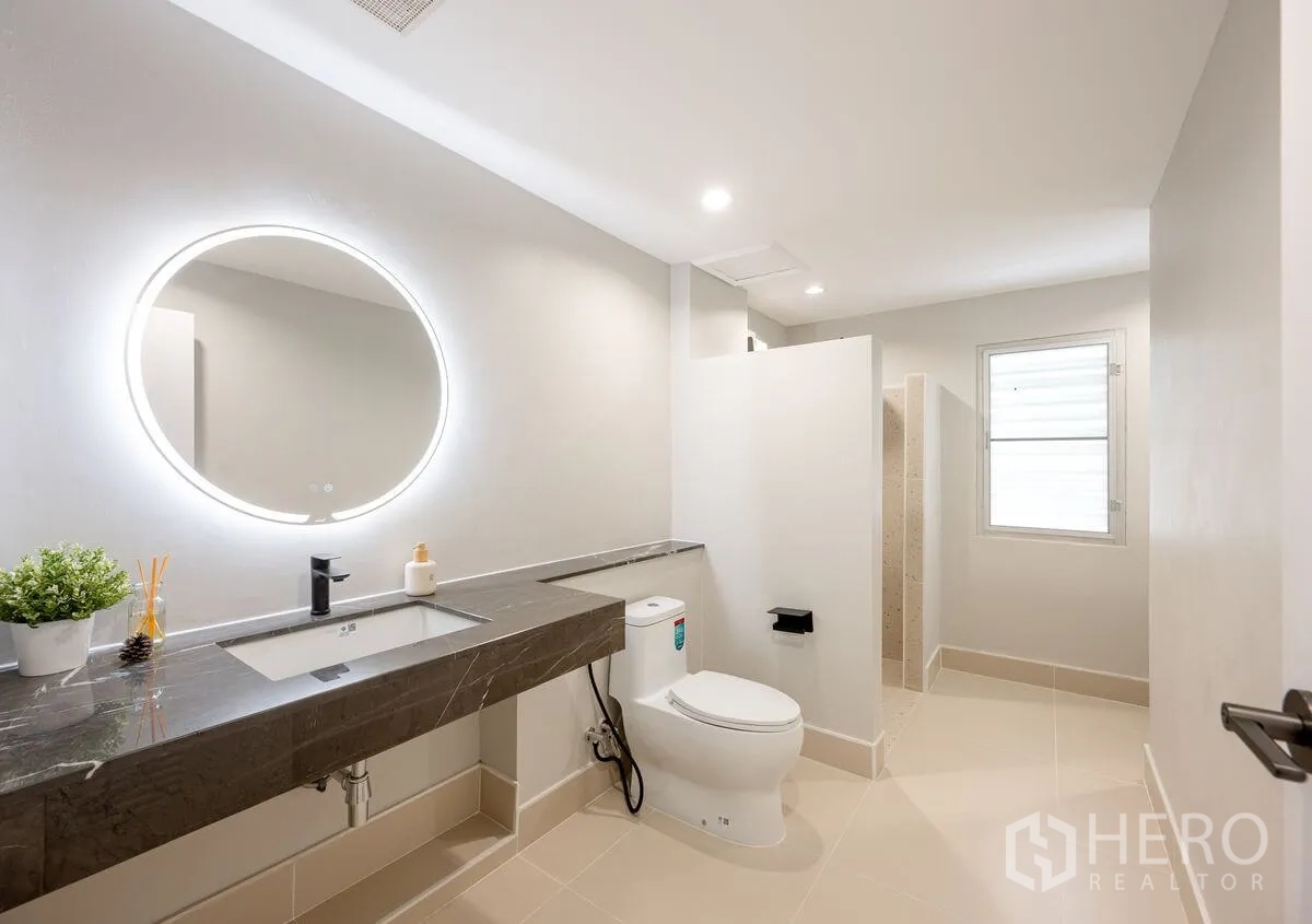 Detached House for rent or sale in Chatuchak, Bangkok - Modern bathroom with long stone vanity, backlit round mirror, toilet, and walk-in shower.