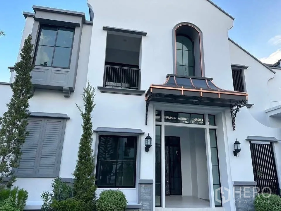 Detached House for sale in Taling Chan, Bangkok - Front facade of a two-storey white detached house with arched window, copper canopy and manicured shrubs.