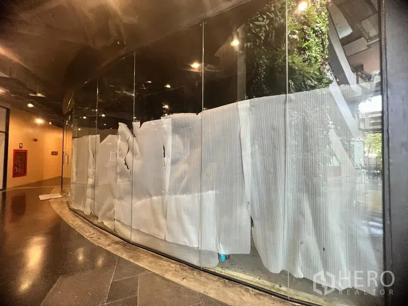 零售空间 出租 - 走廊一侧的弧形落地玻璃店面，玻璃覆盖临时遮挡，室内灯光温暖明亮。