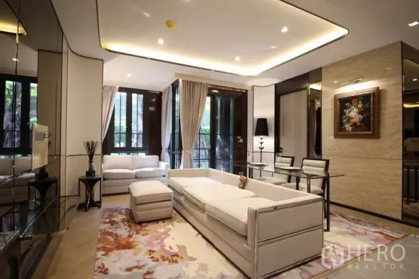 Condominium for rent in Watthana, Bangkok - Elegant living room with white sofas, recessed lighting, large windows and glass dining table overlooking greenery.
