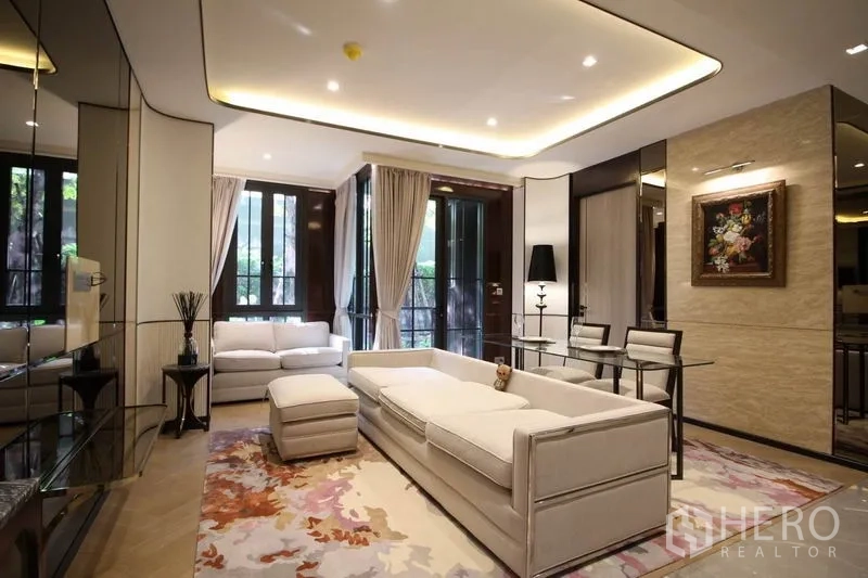 Condominium for rent in Watthana, Bangkok - Elegant living room with white sofas, recessed lighting, large windows and glass dining table overlooking greenery.