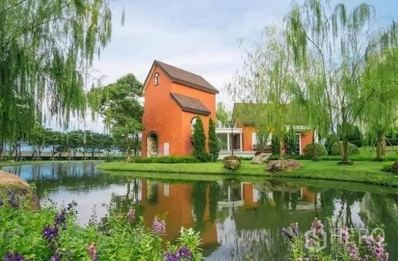 Detached House for rent in Bang Bo, Samut Prakan - Manicured lakeside park with an orange villa, willow trees and a reflective pond.