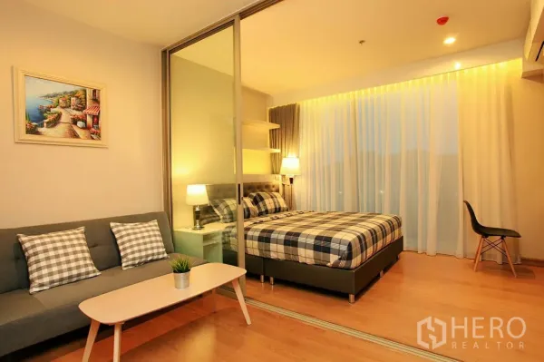 Condominium for rent or sale in Si Racha, Chon Buri - Warm-lit living area with gray sofa and coffee table beside a bedroom behind sliding glass with plaid bedding and sheer curtains.