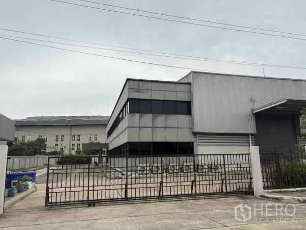 Factory for rent or sale in Samut Prakan - Gated entrance with metal fence facing the office and warehouse facade.