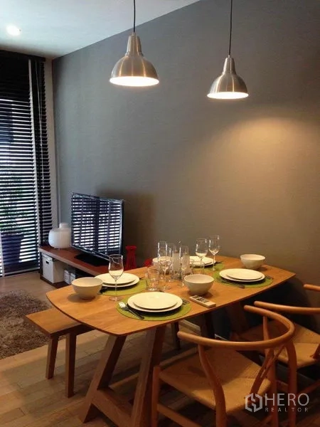 公寓 出租在Watthana, Bangkok - 温馨用餐区，木质餐桌摆放四人餐具，吊灯照明，电视柜与通向阳台的百叶窗。