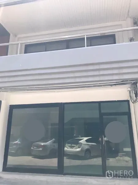 零售空间 出租在Watthana, Bangkok - 一层店面外观，黑色框架落地玻璃滑门，上方有阳台，玻璃倒映停放车辆。