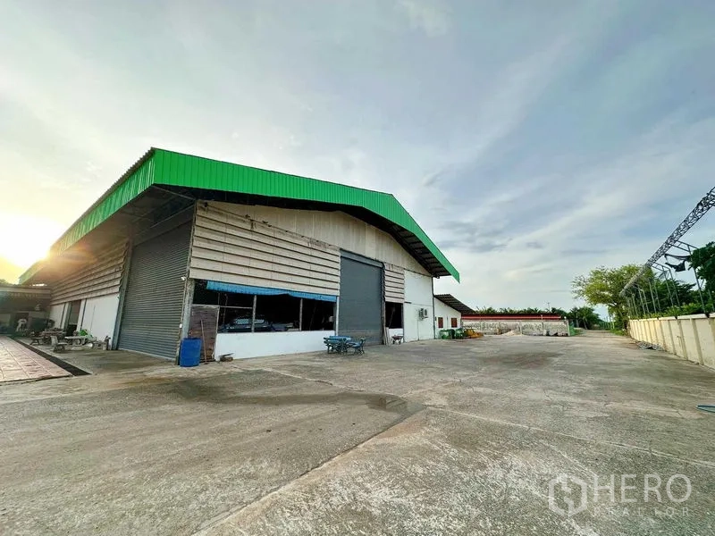 Warehouse for rent in Sai Noi, Nonthaburi - Wide exterior of green-roof warehouse with large concrete yard and roller shutter at sunset.