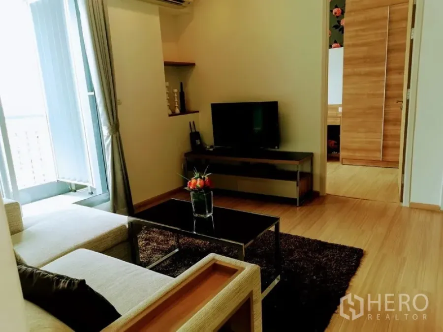 Condominium for rent in Khlong Toei, Bangkok - Bright living room with L-shaped sofa, glass coffee table, TV console and balcony access on wood flooring.