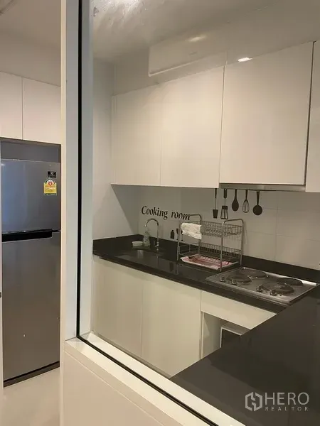 Condominium for rent or sale in Watthana, Bangkok - Modern separated kitchen with white cabinetry, black countertops, cooktop and stainless refrigerator.