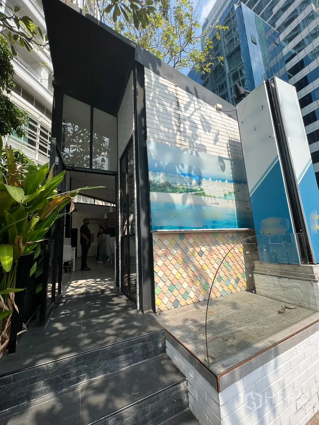 零售空间 出租在Pathum Wan, Bangkok - 现代零售店入口，白色砖墙与玻璃立面，树荫与城市高楼相映。