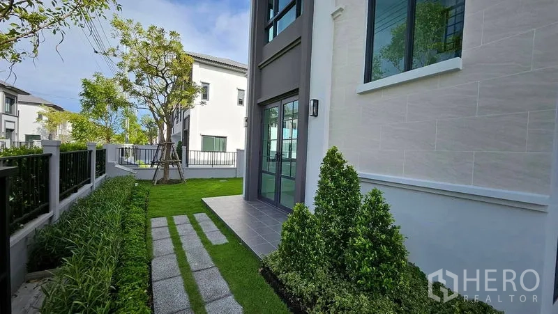 Detached House for sale in Samut Prakan - Side garden with stepping stones, shrubs, and glass door along the home’s contemporary facade.