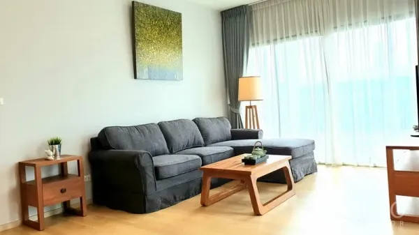 Condominium for rent or sale in Watthana, Bangkok - Bright living room with gray L-shaped sofa, wooden coffee table, and floor-to-ceiling sheer curtains.