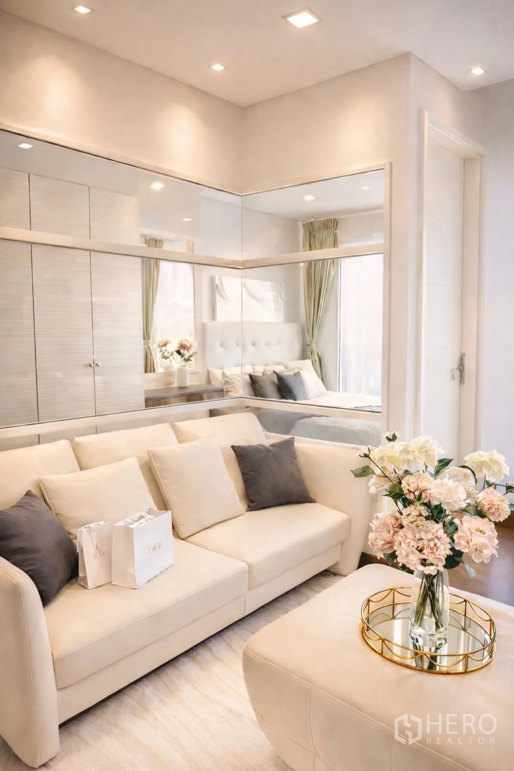 Condominium for rent or sale in Watthana, Bangkok - Bright living area with cream sofa, mirrored wall, and flowers beside a glass partition to the bedroom.