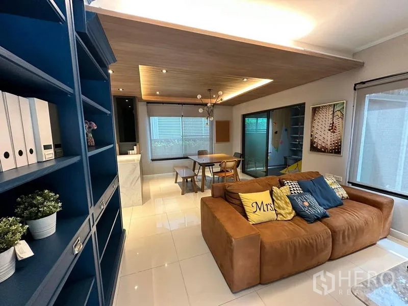 Detached House for rent in Bang Phli, Samut Prakan - Open-plan living and dining area with brown sofa, blue built-in shelves, wood feature ceiling and large windows.