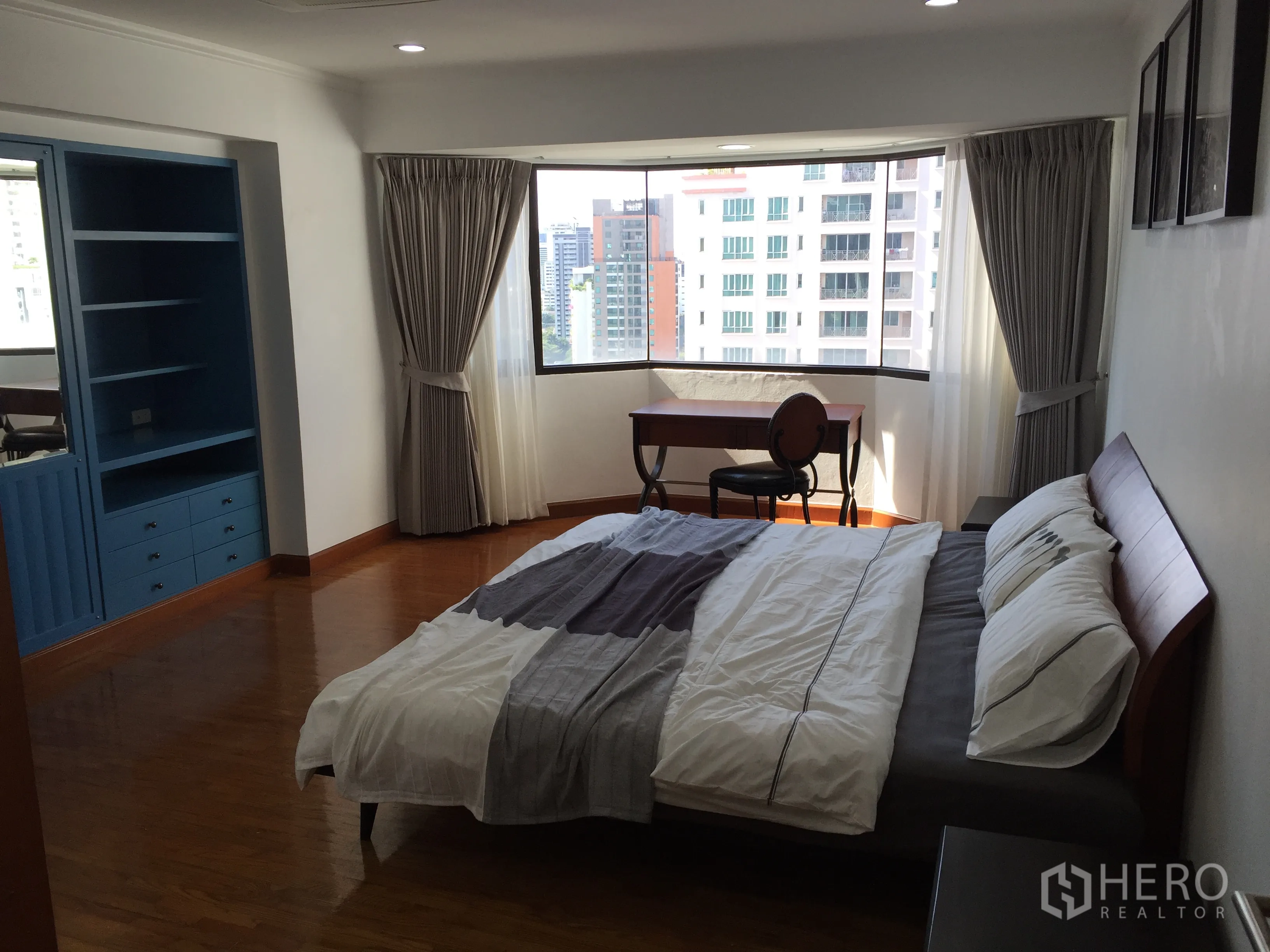 Condominium for sale in Watthana, Bangkok - Primary bedroom with bay window city view, blue built-in shelving, wood flooring and a desk by the window.