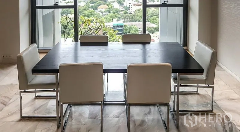 Condominium for rent or sale in Sathon, Bangkok - Modern dining area with six white chairs around a dark table and floor-to-ceiling windows overlooking greenery and city views.