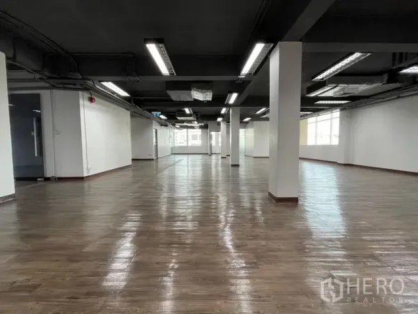 办公空间 出租在Ratchathewi, Bangkok - 开放式办公室楼层，木纹地面、黑色外露天花、白色立柱和明亮灯带，尽头为大窗户。