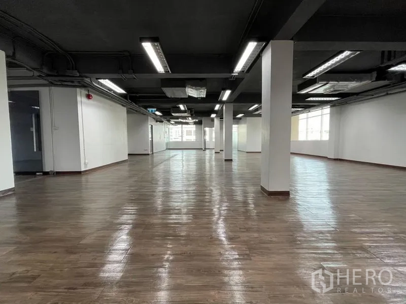 办公空间 出租在Ratchathewi, Bangkok - 开放式办公室楼层，木纹地面、黑色外露天花、白色立柱和明亮灯带，尽头为大窗户。